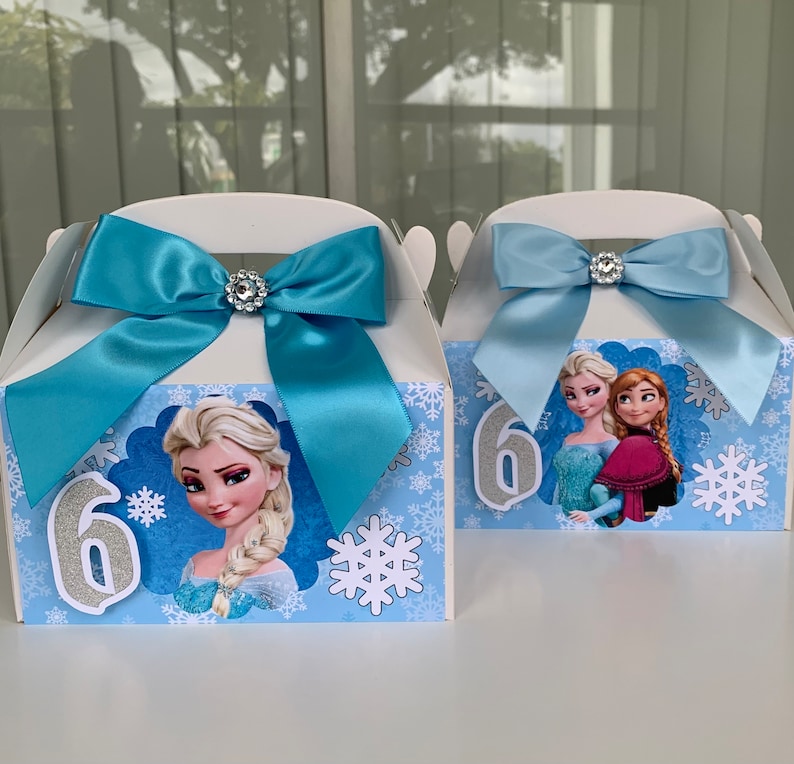 K&ouml;nnte beinhalten: Zwei wei&szlig;e Geschenkboxen mit hellblauen Schleifen und Strassverzierungen. Jede Box zeigt Charaktere aus dem Film "Die Eisk&ouml;nigin", Schneeflocken und die Zahl 6 in silbernem Glitzer.