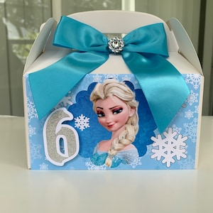 K&ouml;nnte beinhalten: Wei&szlig;e Geschenkbox mit hellblauer Schleife und Strassverzierung. Die Box zeigt ein Bild von Elsa aus Frozen, Schneeflocken und die silberne Zahl 6. Ideal f&uuml;r eine Geburtstagsfeier.