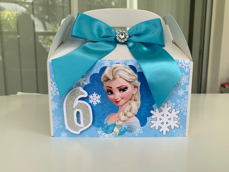 K&ouml;nnte beinhalten: Wei&szlig;e Geschenkbox mit hellblauer Schleife und Strassverzierung. Die Box zeigt ein Bild von Elsa aus Frozen, Schneeflocken und die silberne Zahl 6. Ideal f&uuml;r eine Geburtstagsfeier.