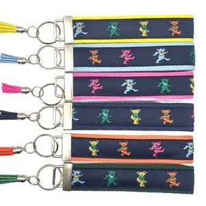 May include: Set of seven keychains with colorful dancing bear designs on navy blue fabric. Each keychain has a silver key ring and a tassel in a different color: yellow, light blue, pink, black, orange, green, and dark green.