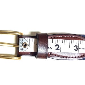 Puede incluir: Un cintur&oacute;n de cuero marr&oacute;n con una hebilla dorada y una correa tejida azul y blanca. La correa tiene una cinta m&eacute;trica con los n&uacute;meros 2, 3 y 4 visibles. El texto "REINFORCED FIBERGL" es visible en la correa.