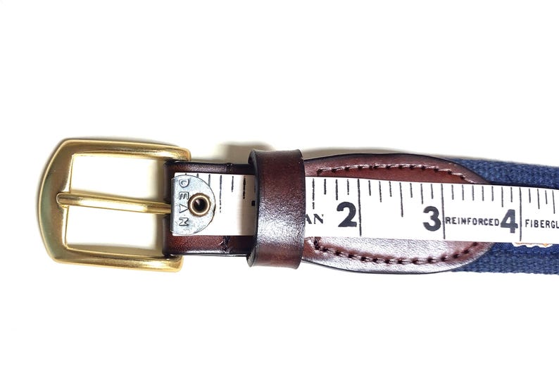 Puede incluir: Un cintur&oacute;n de cuero marr&oacute;n con una hebilla dorada y una correa tejida azul y blanca. La correa tiene una cinta m&eacute;trica con los n&uacute;meros 2, 3 y 4 visibles. El texto "REINFORCED FIBERGL" es visible en la correa.