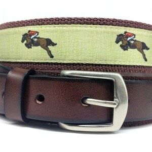 May include: A brown leather belt with a silver buckle. The belt has a green and brown striped fabric band with a repeating pattern of a rider on a horse.