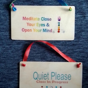 May include: Two wooden signs with colourful text and embellishments. The top sign says "Meditate Close Your Eyes & Open Your Mind" with a rainbow chakra symbol. The bottom sign says "Quiet Please Class In Progress".