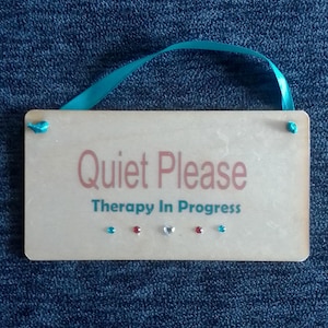 May include: A wooden sign with the text "Quiet Please Therapy In Progress" in pink and green lettering. The sign has a turquoise ribbon for hanging and is decorated with turquoise, red, and silver rhinestones.