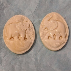 May include: Two oval-shaped, cream-colored buttons with a carved design of elephants. The elephants are facing each other and have intricate details.