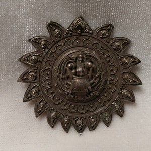 May include: A silver-toned metal brooch with a circular design featuring a central figure and intricate details. The brooch has a starburst shape with multiple points.