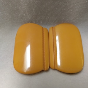 May include: A pair of vintage yellow plastic belt buckles. The buckles are smooth and shiny, with a slight curve to them.