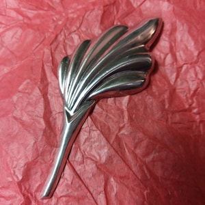 May include: A polished, silver-toned metal decorative piece, shaped like a stylized feather or leaf. The object has a long, slender stem and a fan-like top with multiple curved ridges. It rests on a textured, red surface.