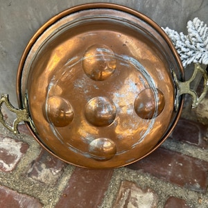 May include: A vintage copper pan with brass handles. The pan has a round shape with five raised circular details on the interior. The copper has a warm, reddish-brown hue with some areas of patina. The handles are ornate and add a decorative touch.