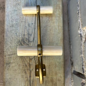 May include: A brass-toned metal latch with two ivory-colored wooden handles, mounted on a rectangular wooden board with a distressed finish. The latch is designed for a door or cabinet.