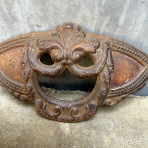 May include: A vintage, ornate, cast iron door pull with a ring handle. The pull is rusty brown and has a decorative design.