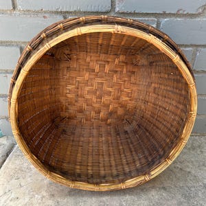 May include: A large, round, woven basket made of natural brown wicker. The basket has a wide, open top and a slightly rounded bottom. The basket is made of a tightly woven pattern of thin strips of wicker.