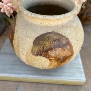 May include: A round, speckled ceramic pot with a wide opening and a thick, textured rim. The exterior features a light beige, sandy base color with large, irregular patches of dark brown, glossy glaze applied in a circular, organic pattern.
