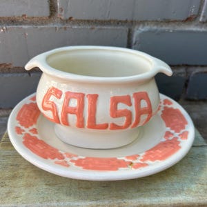 May include: Cream-colored ceramic salsa bowl and saucer set. The bowl has two small handles and the word "SALSA" in coral-colored letters. The saucer features a coral-colored geometric pattern around the rim.