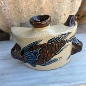 May include: A small, semi-circular ceramic vessel with a beige base and a dark brown glazed rim and spout. The front features a relief design of a fish with blue fins and a textured, dark brown body. Two small handles extend from the sides.