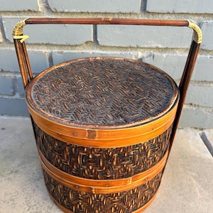 May include: A round, woven bamboo basket with a lid and a handle. The basket is primarily brown with lighter brown accents and a woven pattern. The handle is made of a darker wood and is attached with gold-colored metal.