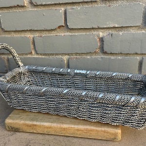 May include: A rectangular metal wire basket with two arched handles. The basket is made of woven silver-colored wire and is supported by a wooden base. The basket is placed in front of a gray brick wall.