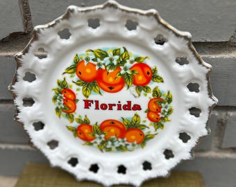 May include: White decorative plate with a gold rim and a scalloped edge. The plate features a colorful illustration of oranges, green leaves, and the word "Florida".