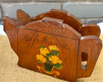 May include: Wooden teapot-shaped napkin holder with a painted design of yellow flowers in a green pot with a yellow ribbon. The holder is painted with the word "remarkable" on the front.