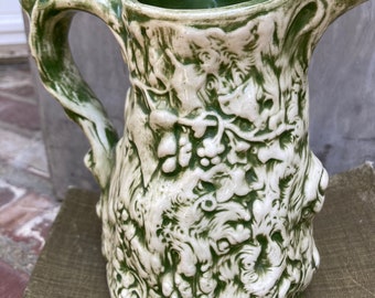 May include: A green and white ceramic pitcher shaped like a tree trunk with a detailed, textured design. The pitcher has a wide, rounded base and a handle that curves up and over the top.