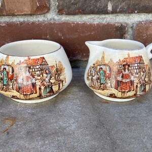 May include: A vintage sugar bowl and creamer set with a cream-colored background and a colorful scene of people in a town square. The scene includes a horse-drawn carriage, a building with a pointed roof, and several people in period clothing.