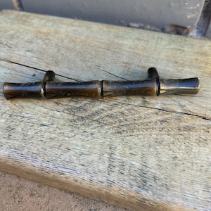 May include: A dark brown metal cabinet pull with a bamboo-like design. The hardware is made of metal and has a vintage or antique appearance. The pull is lying on a weathered wooden surface.