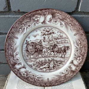 May include: A decorative plate with a brown and white transferware design. The plate features a rural scene with a horse-drawn carriage and floral border. The plate is resting on an open book.