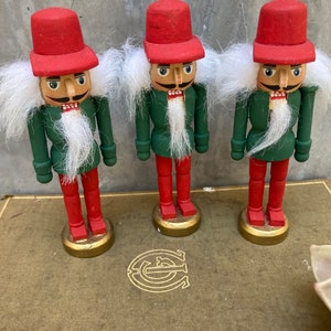 May include: Three wooden nutcrackers with red hats, green jackets, and red pants stand on a gold-colored base. Each has a white beard and mustache. The nutcrackers are arranged on a vintage book.