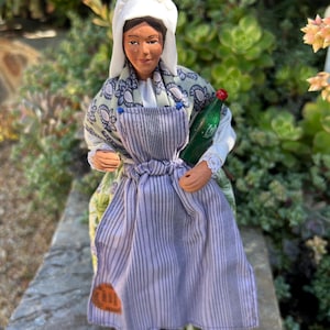 May include: A ceramic figurine of a woman wearing a white cap, a green and blue patterned shawl, a purple and white striped apron, and holding a green glass bottle.