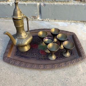 May include: A brass tray with a floral design and six small brass cups, and a brass teapot with a spout and a lid.
