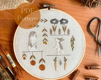 May include: A white embroidery hoop with a brown wooden frame. The hoop contains a hand-stitched design of two rabbits, flowers, and carrots. The design is done in shades of brown, green, and white. The hoop is surrounded by brown wooden clothespins and brown yarn spools.