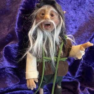 May include: A handmade gnome doll with a white beard and brown and green clothing. The gnome has a surprised expression on its face.