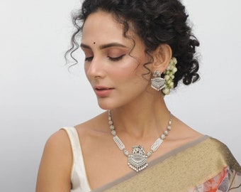 May include: A woman with curly hair wears a silver necklace with a pearl and ornate pendant. She also wears silver earrings with a similar design. The necklace and earrings are on a white background.
