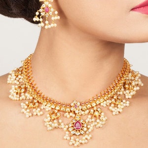 Puede incluir: Un collar de oro con un colgante de piedra rosa y detalles de perlas. El collar está adornado con detalles intrincados y tiene un diseño tradicional indio.