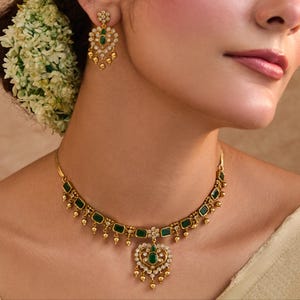 Puede incluir: Juego de joyas doradas con collar y pendientes. El collar tiene piedras rectangulares verde esmeralda y un colgante en forma de corazón con una piedra verde central. Los pendientes reflejan el diseño con una piedra verde central.