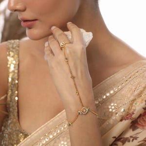 May include: A gold-toned hand chain with a ring and bracelet design. The ring features a floral design with a central white stone and surrounding smaller stones. The chain is adorned with small green and clear stones and star-shaped accents.
