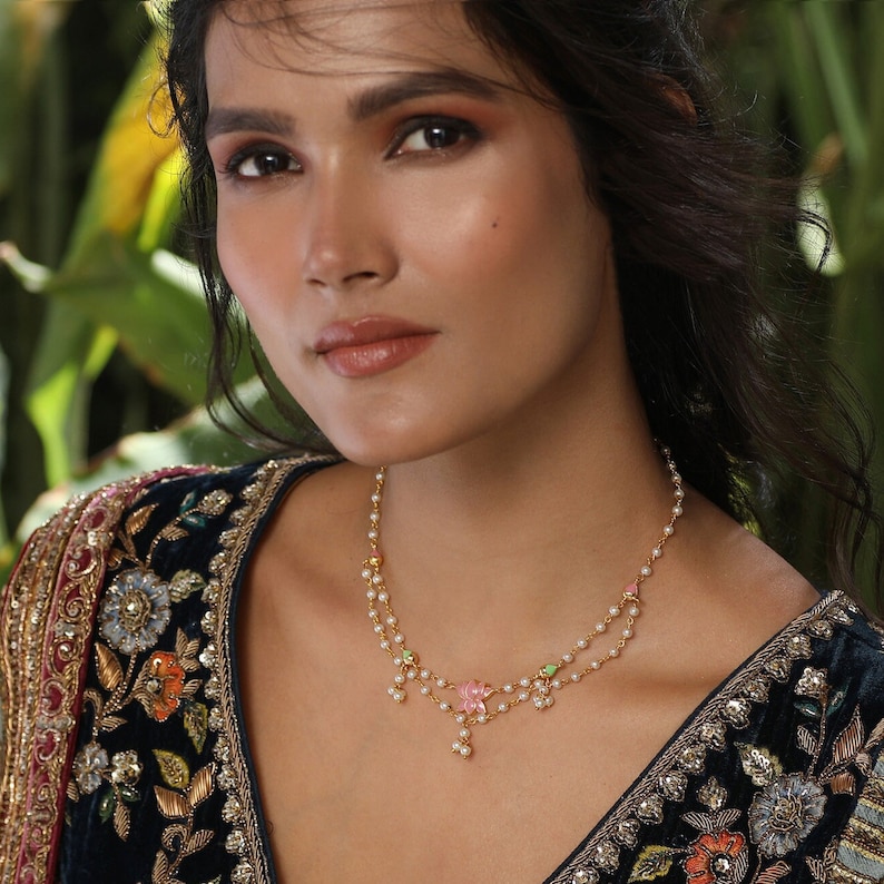 May include: A gold necklace with a delicate pearl chain and a pink flower pendant. The necklace is layered with a second chain that has a green flower pendant. The necklace is worn by a woman with dark hair and makeup.