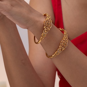 May include: Two gold bangles featuring detailed floral and scroll patterns. The bangles are embellished with small red, green, and clear gemstones. The jewellery is shown on a person's wrist, set against a red backdrop.