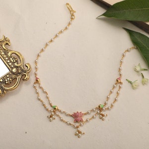 May include: A gold chain necklace with a pink and green enamel lotus flower pendant. The necklace is made of small white beads and has a hook clasp.