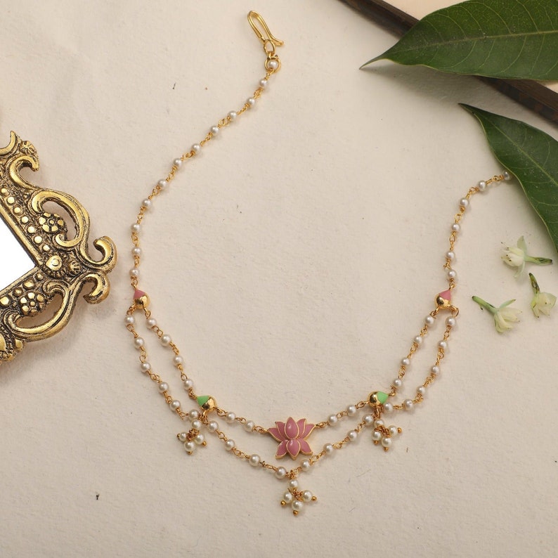 May include: A gold chain necklace with a pink and green enamel lotus flower pendant. The necklace is made of small white beads and has a hook clasp.