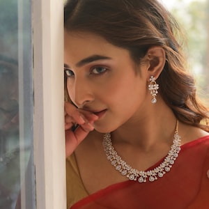 Puede incluir: Una mujer lleva un sari rojo y dorado con un collar y pendientes de diamantes. El collar presenta un diseño floral con piedras colgantes en forma de lágrima.