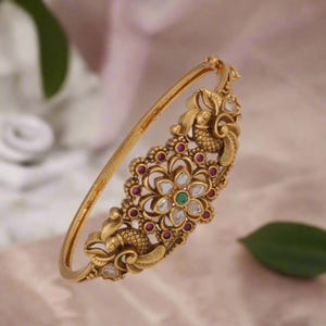 Puede incluir: Un brazalete de oro con intrincados diseños florales y de pájaros. El brazalete está adornado con piedras preciosas transparentes y verdes.