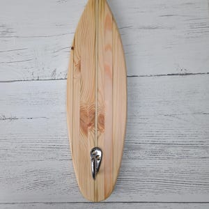 May include: A wooden surfboard-shaped wall hook. The hook is made of light-colored wood with a natural grain pattern. A silver-colored metal hook is attached to the bottom. The surfboard is approximately 30 inches long.