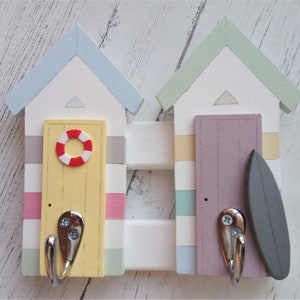 May include: A wooden beach hut-shaped coat rack with two hooks. The left hut is painted yellow, blue, pink, and gray with a red and white life preserver on the door. The right hut is painted purple, green, blue, and yellow with a gray surfboard on the door.