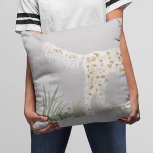 May include: A throw pillow with a watercolor illustration of a white and brown spotted dog with a long tail, standing in a field of green grass with small pink flowers. The background is a light gray color.