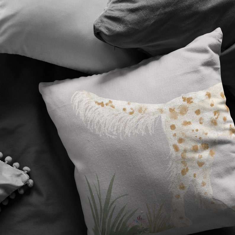 May include: A white throw pillow with a brown and white spotted dog design. The dog is lying down with its tail curled up. The pillow is on a gray bed with other pillows.