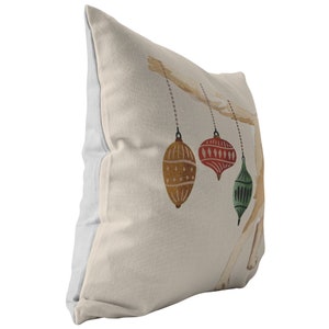 May include: A beige throw pillow with a white border features a watercolor illustration of three Christmas ornaments hanging from a branch. The ornaments are red, green, and gold.