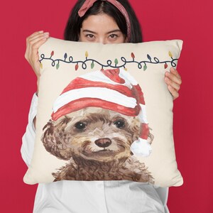 May include: A cream-colored throw pillow with a watercolor illustration of a brown and white dog wearing a red and white Santa hat. The pillow has a string of colorful lights printed on it.