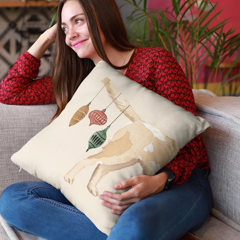 May include: A beige throw pillow with a watercolor illustration of a dog with three hanging ornaments. The ornaments are green, red, and brown.